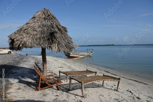 Sillas y tumbonas rusticas debajo de una sombrilla de paja, mar tranquilo y cielo despejado, barco anclado en la Playa de Bomani en Tanzania, África del Este.