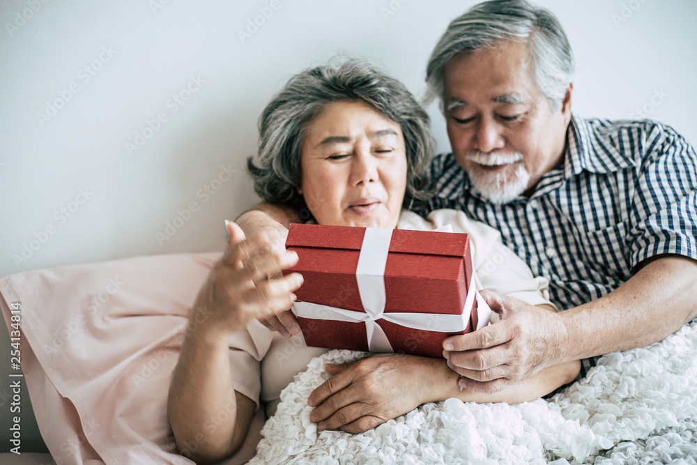 Smiling senior husband making surprise giving gift box to his wife in bedroom Smiling senior husband making surprise giving gift box to his wife in bedroom