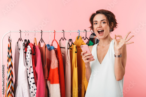 Happy woman stylist posing isolated over pink wall background near a lot of clothes using mobile phone. photo