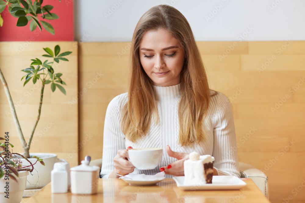 Photo of relaxed light haired female drinks hot coffee or cappucino with delicious dessert, sits in cosy cafeteria, waits for friends, enjoys spare time during weekend. People, lifestyle, rest concept Photo of relaxed light haired female drinks hot coffee or cappucino with delicious dessert, sits in cosy cafeteria, waits for friends, enjoys spare time during weekend. People, lifestyle, rest concept