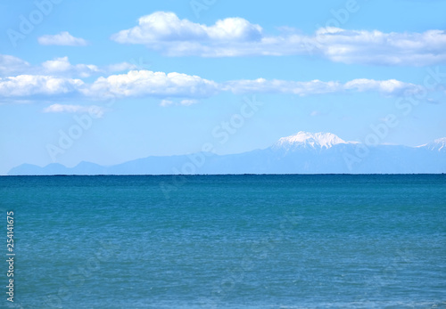 High mountains silhouettes with white snow cap on top far away on horizon in light haze after the sea under blue sky with white cumulus clouds