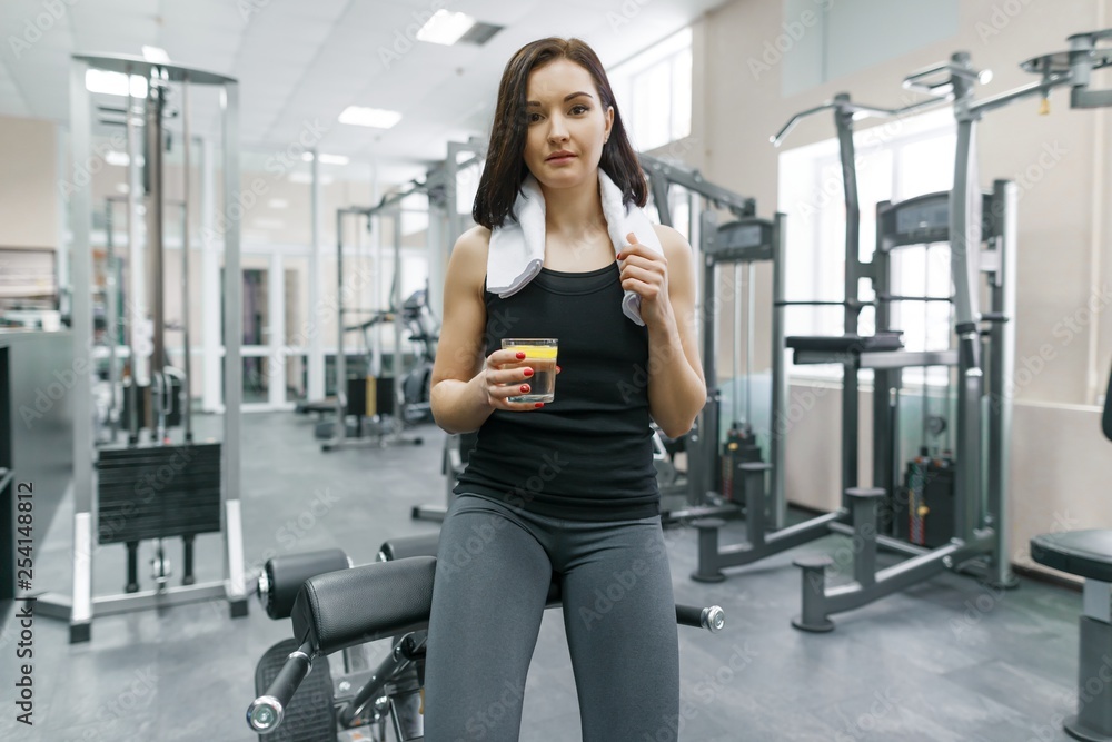 Portrait of young beautiful fitness woman in sportswear with towel, with glass of water and lemon, after fitness classes drinking water in gym. Healthy lifestyle and nutrition