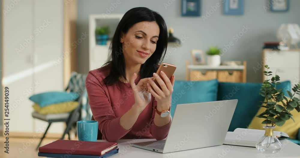 Young good looking brunette woman sitting at the table with a laptop computer and texting message on the smartphone in the living room. Young good looking brunette woman sitting at the table with a laptop computer and texting message on the smartphone in the living room.
