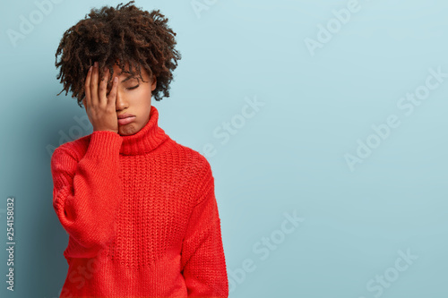 Fotografia Discontent tired dark skinned woman covers face with hand, has tired expression, wears red sweater, isolated over blue wall with empty space for your promotion