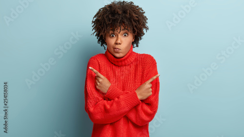 Fotografie Confused black woman points sideways, suggests looking in different sides, purse