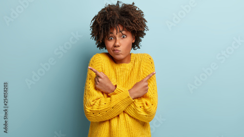 Puzzled confused woman has hesitant expression, curly hairstyle, crosses hands over chest, points right and left in different sides, wears yellow sweater, being indifferent, models over blue wall