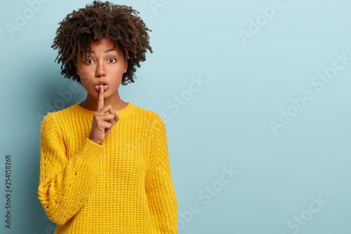 Studio shot of quiet dark skinned lady keeps fore finger over lips  makes silence gesture  gossips with friend  wears yellow sweater  says hush  isolated over blue studio wall with blank space