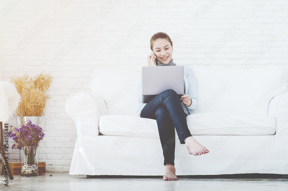 Beautiful Asian woman is smiling.Asian women work with gray laptops on the sofa in the room in the morning.Orange shopping bag in his hand.Girl in white shirt. Beautiful Asian woman is smiling.Asian women work with gray laptops on the sofa in the room in the morning.Orange shopping bag in his hand.Girl in white shirt.
