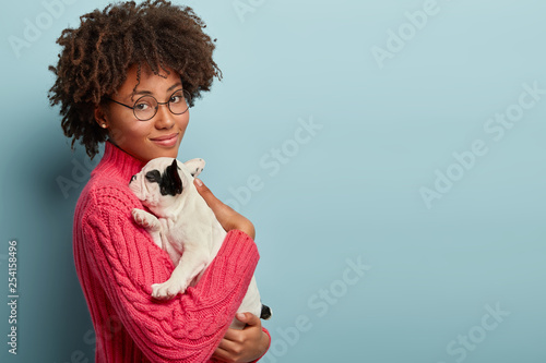Sideways shot of calm tender woman holds small sleepy puppy closely to chest, snuggles french bulldog wears round spectacles, carries dog for having outdoor walk. Friendship between people and animals