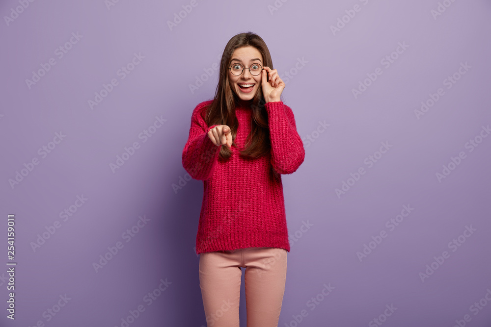 Teenage girl with happy expression indicates directly at camera, points with fore finger, greets friend, wears optical glasses and red jumper, isolated over purple studio wall, invites guests, hints Teenage girl with happy expression indicates directly at camera, points with fore finger, greets friend, wears optical glasses and red jumper, isolated over purple studio wall, invites guests, hints