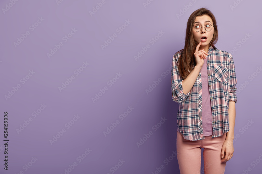 Thoughtful puzzled woman opens mouth with surprisement, focused aside, tries to overhear rumors, wears stylish checkered shirt, optical glasses, isolated over purple wall with mock up space. Thoughtful puzzled woman opens mouth with surprisement, focused aside, tries to overhear rumors, wears stylish checkered shirt, optical glasses, isolated over purple wall with mock up space.