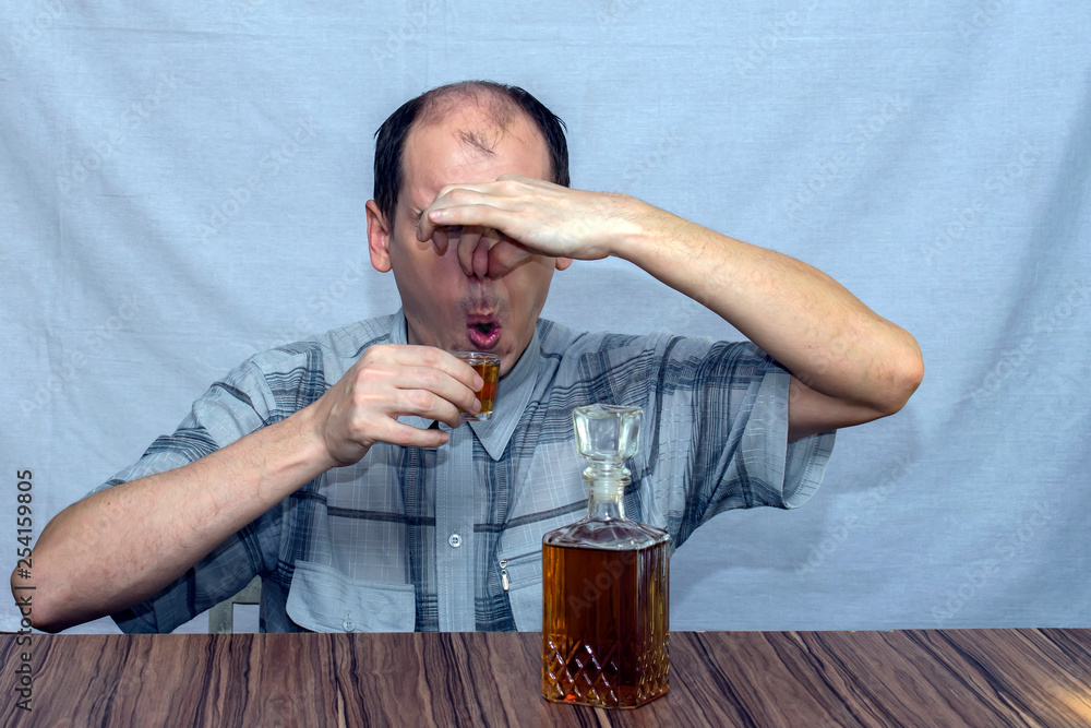 A man sits at the table and wants to drink alcohol. At the same time he squeezed his nose with his fingers and closed his eyes. Before him stands a bottle of alcohol. A man sits at the table and wants to drink alcohol. At the same time he squeezed his nose with his fingers and closed his eyes. Before him stands a bottle of alcohol.