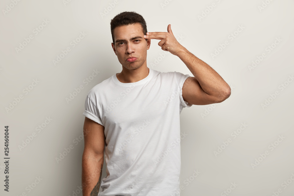 Horizontal shot of serious tired attractive guy shoots in temple with finger, demonstrates suicide gesture, wears white t shirt, fed up of long work, has strong body, isolated over white background. Horizontal shot of serious tired attractive guy shoots in temple with finger, demonstrates suicide gesture, wears white t shirt, fed up of long work, has strong body, isolated over white background.
