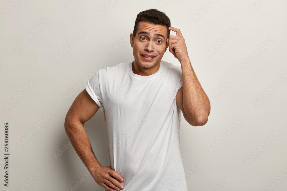 Frustrated dissatisfied young man scratches head, forgot about home duties, keeps hand on waist, stares at camera in despair, wears white t shirt, has amnesia or bad memory. Hmm, let me think Frustrated dissatisfied young man scratches head, forgot about home duties, keeps hand on waist, stares at camera in despair, wears white t shirt, has amnesia or bad memory. Hmm, let me think