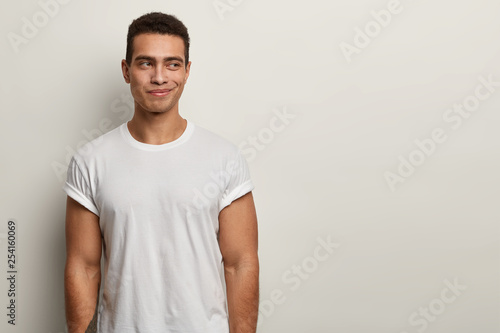 Photo of satisfied smiling man with thoughtful expression, muscular body, wears white t shirt in one tone with background, thinks about how spend next weekend, has tattooed arm. Youth concept
