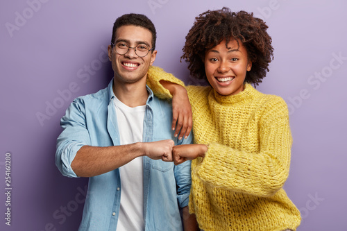 Young diverse couple give fist bump, agree to bring plan to life, smile broadly, have mixed race relationships, have good friendly partners, isolated over purple wall. I agree with you concept photo