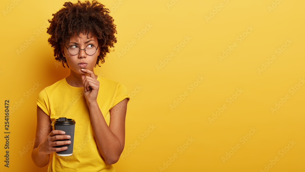 Studio shot of thoughtful African American woman holds chin, contemplates about something with angry serious expression, holds paper cup of hot drink, has coffee break, isolated on yellow wall