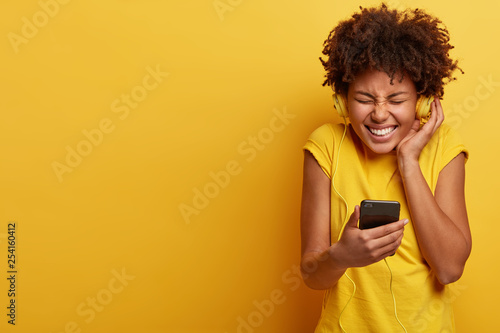 Photo of joyful teenager squints face, has broad smile, has white teeth, keeps hands on headphones, listens music with high volume, holds modern cell phone, chills with good vibes. Hobby concept