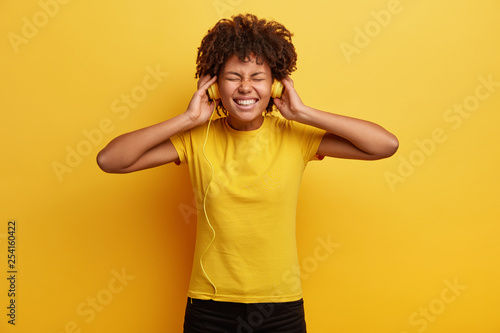 Overjoyed amused African American woman enjoys leisure time, keeps both hands on stereo headphones, listens favourite song, has toothy smile, dressed in bright yellow t shirt being real lover of music