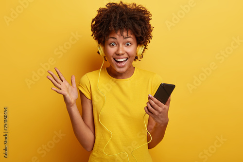 Photo of laughing impressed lovely woman looks happily at camera, raises palm, holds cell phone, ready to go for walk with music, likes yellow colour, looks directly at camera with satisfaction