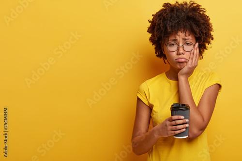 Upset dark skinned female feels sleepy and discontent to wake up early in morning, holds paper cup of coffee, wears transparent eyewear and yellow t shirt, keeps hand on cheek, being fatigue
