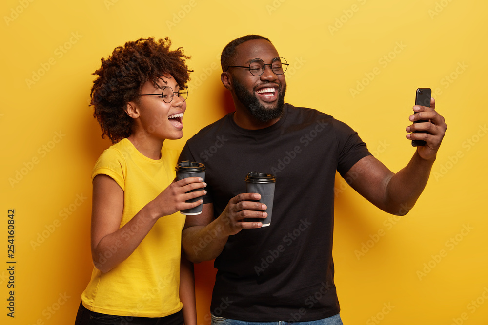 Indoor shot of happy ethnic girlfriend and boyfriend take selfie on modern gadget, laugh as being in good mood, drink coffee during spare time, express positive emotions, isolated on yellow wall Indoor shot of happy ethnic girlfriend and boyfriend take selfie on modern gadget, laugh as being in good mood, drink coffee during spare time, express positive emotions, isolated on yellow wall