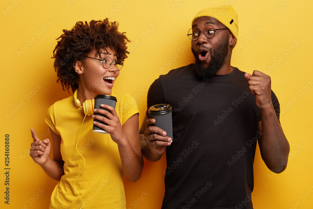 Happy joyful young woman and man look surprisingly positively, gesture actively while dance with loud music, drink takeaway coffee, celebrate successful day, isolated over yellow background. Happy joyful young woman and man look surprisingly positively, gesture actively while dance with loud music, drink takeaway coffee, celebrate successful day, isolated over yellow background.