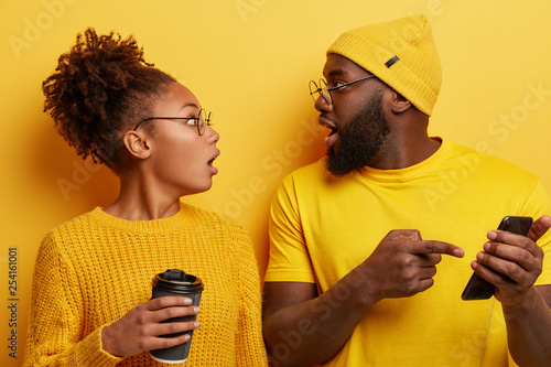 Shocked black woman and man impressed while scroll social media page. Emotioanl lady with crisp hair hears shocking newsfrom friend who indicates at cell phone screen, holds paper cup of beverage photo