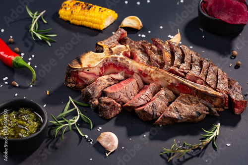 porterhouse t-bone steak is grilled sliced on a piece with grilled corn, sauce, chilli, rosemary, salt, garlic on black background. rustic style
