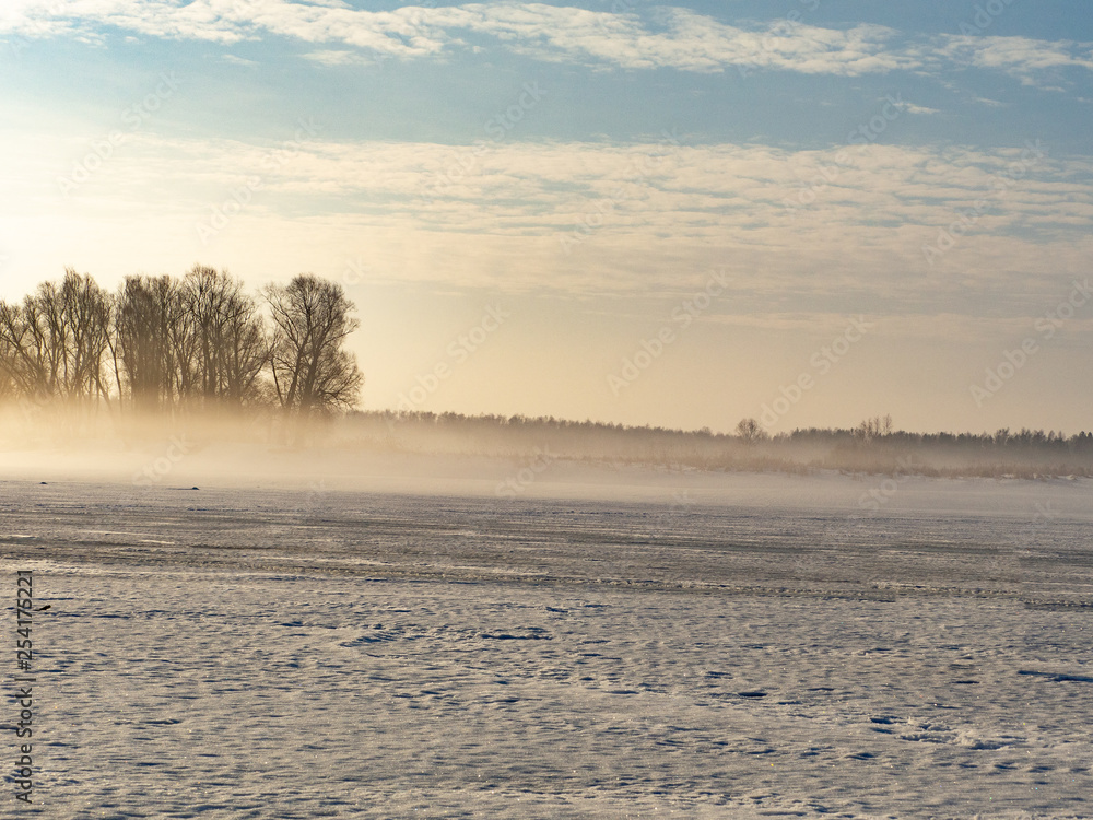 Foggy winter landscape with low sun, snow and river in the ice. Trees grow along the shore.