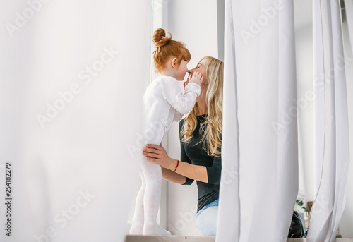 Happy mother kissing little redhead daughter near large window with the white curtains