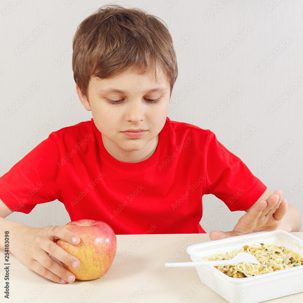 Young boy at the table chooses between instant noodles and apple on a white background Young boy at the table chooses between instant noodles and apple on a white background