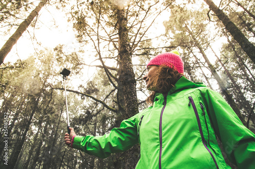 View from below middle aged woman brown curly hair in mountain forest takes selfie with camera among tall trees. Wearing technical clothing wool cap bright colors fluorescent pink green yellow