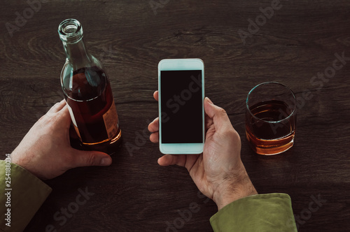 A man holds a mobile phone in his hands. Next on the table is a glass and a bottle of whiskey.