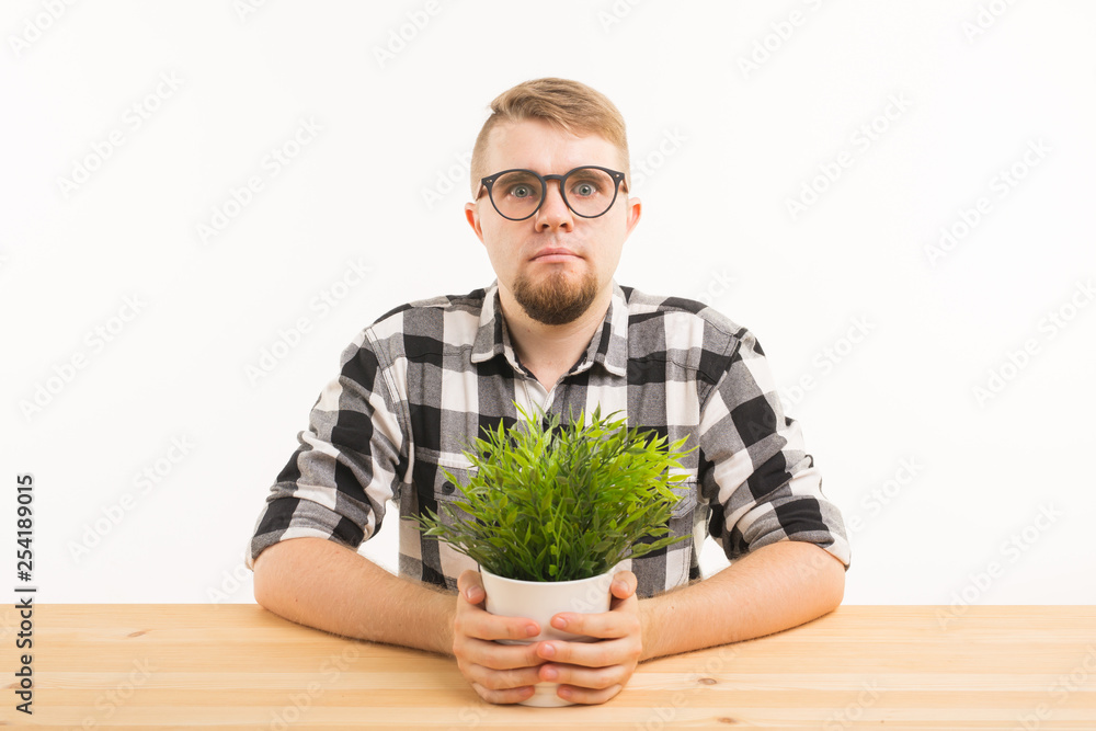 Student, office and people concept - a young grumpy man sitting at the table with a green plant on white background. He looks serious and strange Student, office and people concept - a young grumpy man sitting at the table with a green plant on white background. He looks serious and strange