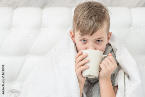 Sick boy with warm scarf sitting on the bed and drink hot tea. Empty space for text