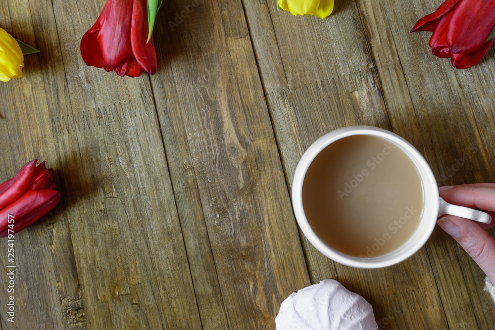 Flat lay of greeting card with flowers tulips and hand keeping white cup of coffee with milk on brown wooden background. Top view. Copy space Flat lay of greeting card with flowers tulips and hand keeping white cup of coffee with milk on brown wooden background. Top view. Copy space