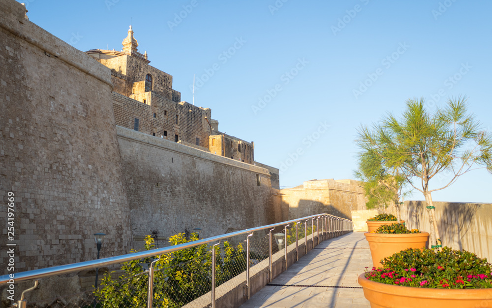 Outside view on the citadel of Victoria from the castle wall covered with flowers in pot and handrail surrounded by sandstone wall in Gozo on Malta.