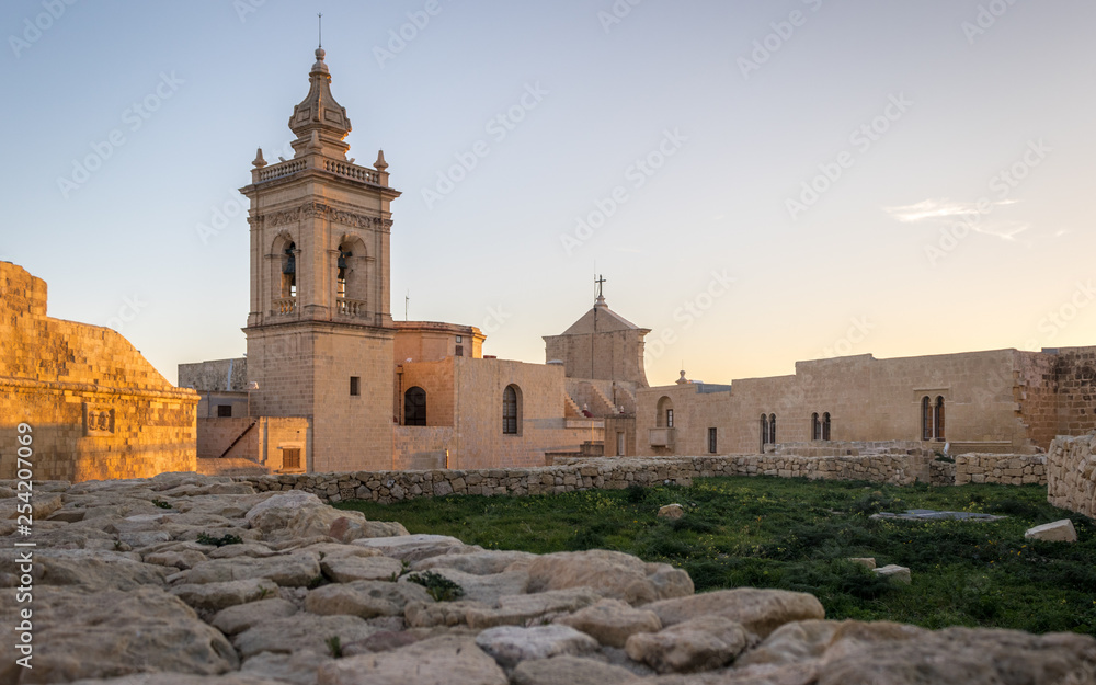 Side view on the old, historical St. Joseph's Chapel inside the Citadel of Victoria surrounded by antique ruins, walls with grass field and sandstones in the foreground on Gozo, Malta.