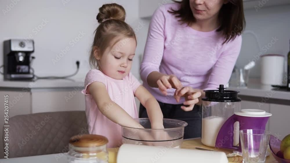 Happy young girl with her mother making dough. Mother and daughter baking in kitchen. Girl trying to mix the dough with hands Happy young girl with her mother making dough. Mother and daughter baking in kitchen. Girl trying to mix the dough with hands