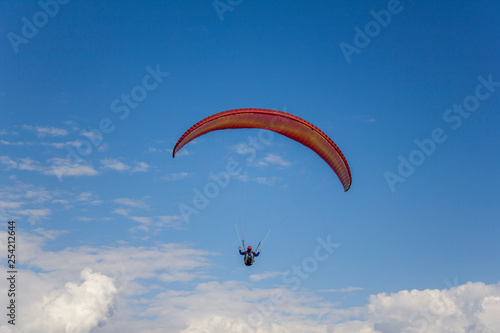 paraglider on a red parachute flies in a clear blue sky with white clouds aerial view
