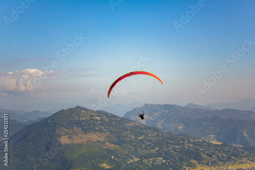 paraglider on a red parachute flies over a green mountain valley under a clear blue sky aerial view