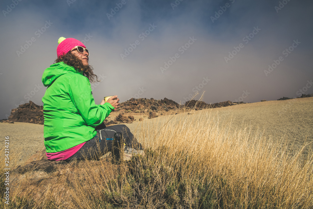Curly brunette woman with wool cap drink coffee sitting on the rocks after trekking in mountain wearing bright fluorescent clothing Girl relaxing with hot beverage. Sand and blue sky in the background