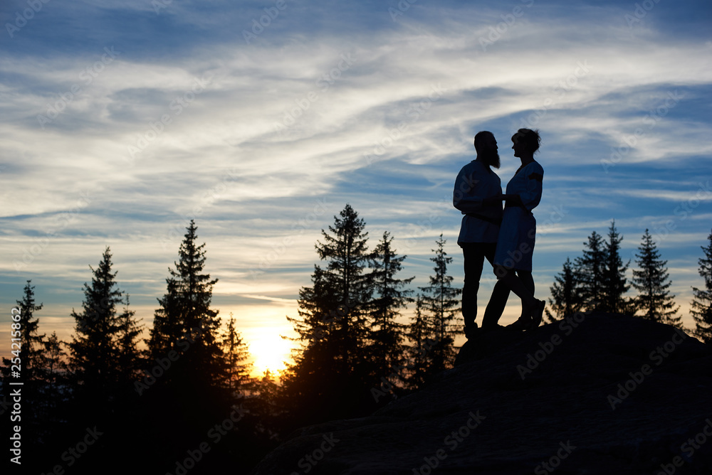 Silhouettes of young couple in love standing on the top of mountain in the evening. Bearded man and happy girl enjoying beautiful sunset, looking to each other. Cloudy sky and forest on background.