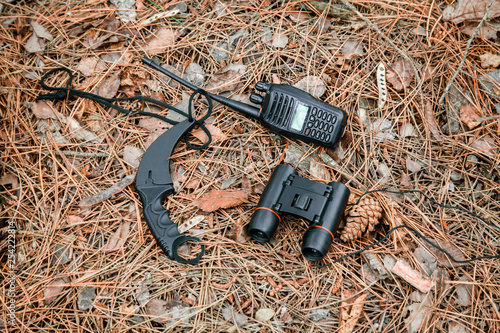 binoculars, radio set and karambit knife on the ground covered with pine needles photo