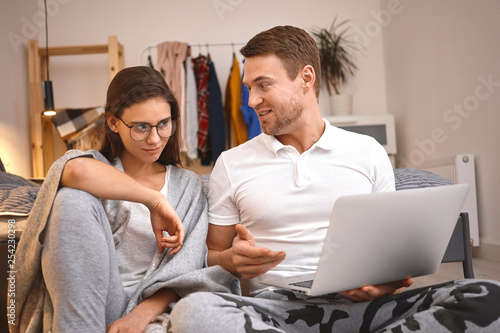 Cute young woman in eyewear sitting next to her husband listening to him talking about prospective internet project. Beautiful Caucasian couple shopping online using generic portable computer