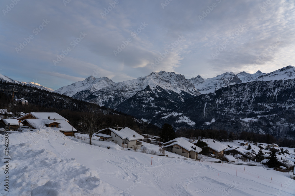 Abendhimmel mit Wolken über Bergdorf im Schweizer Skiort Hohfluh im Hasliberg. Sicht vom Wegesrand über schneebedeckte Piste ins Tal hinunter. 