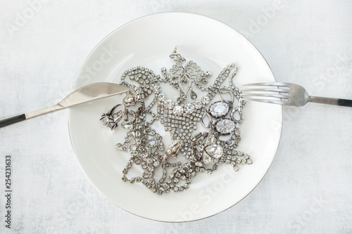 Many sparkling jewelery on a white plate with a knife and fork. The concept of luxury life, wealth, glamor, fashion and weddings