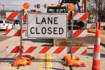 Lane Closed sign at messy construction urban intersection with sand bags traffic cones heavy equipment working and cars stopped at stoplight