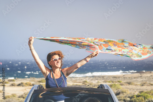 Middle-aged woman waving headscarf out of hood of convertible car. Curly brunette girl holds in hands raised colored pareo, Person having fun in cabriolet, blue sea and sky in the background